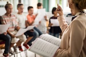 Unrecognizable music teacher with sheet music practicing with choir of children in the classroom.