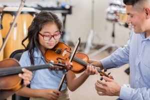 A smiling little girl sits next to her attentive music teacher and plays the violin during music class.  Her teacher adjusts the violin's position as she plays.